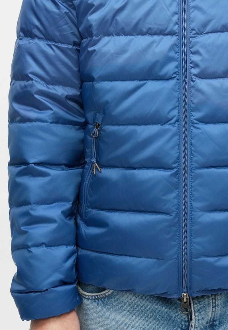 Blue padded jacket with a smooth texture, horizontal quilted design, and a front zipper. Features a zippered side pocket.