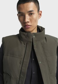 Olivgrüne gepolsterte Weste mit hohem Kragen, Druckknöpfen und Fronttaschen. Sie verfügt über einen Reißverschluss und gerippte Designelemente.