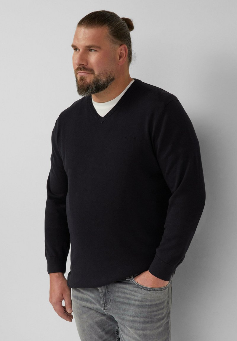 Marineblauer V-Ausschnitt-Pullover aus weichem Baumwollmix, mit langen Ärmeln und gerippten Bündchen, kombiniert mit hellgrauen Jeans.