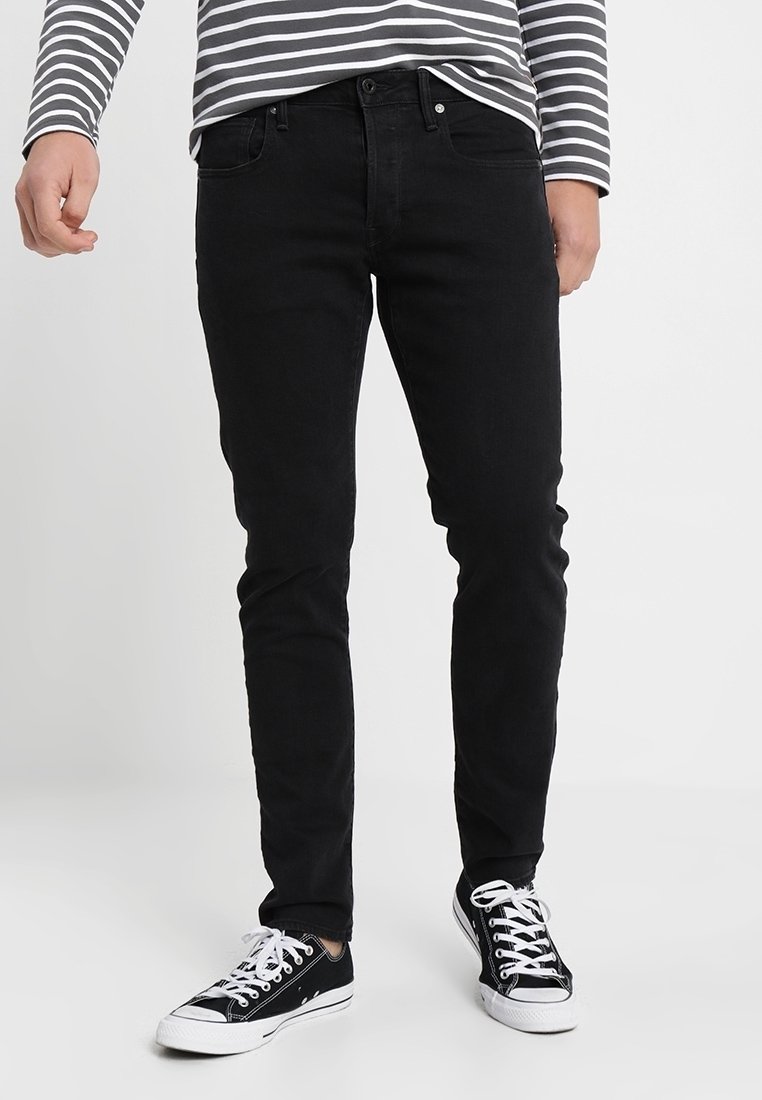 Jeans en denim noir à coupe slim, design cinq poches, couture contrastante, associés à des baskets noir et blanc. Chemise à manches longues avec rayures horizontales.