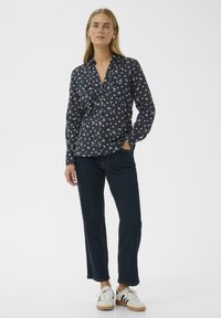 Femme se tenant debout, portant une chemise boutonnière florale marine, un jean droit bleu foncé et des baskets blanches avec des rayures noires.