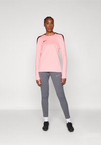 Top sportiv roz cu mâneci lungi și accente negre pe umeri, asortat cu pantaloni gri ajustați. Pantofi sport negri și șosete albe completează ținuta.
