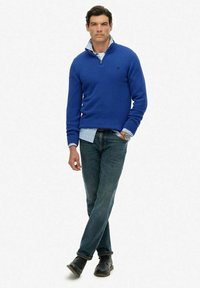 Blauwe gebreide trui met een ritssluiting bij de hals, gedragen over een licht gestreept shirt, gecombineerd met donkere denim jeans en zwarte schoenen. Eenvoudig ontwerp en pasvorm.