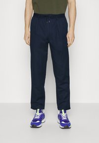 Mörkblå, löst sittande byxor med elastiskt midjeband och dragsko. Bärs med lila träningsskor som har gröna detaljer och vita snören.