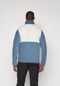 Pullover in pile di colore azzurro chiaro e bianco, con colletto alto e un design a blocchi di colore. Texture liscia con una vestibilità comoda.