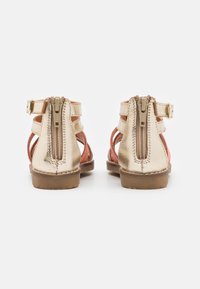 Kulta-korallinväriset metalliset sandaalit, joissa on avoin takapuoli, säädettävä hihna ja vetoketjukiinnitys. Texturoitu pinta ja kestävä kumipohja.