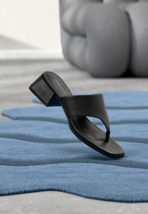 Sandalia negra de tacón con tira ancha sobre superficie azul texturizada, sillón marrón difuminado al fondo.