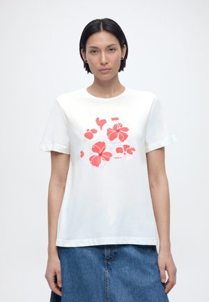 Kobieta o krótkich ciemnych włosach, ubrana w białą koszulkę z czerwono-różowym kwiatowym nadrukiem i niebieską spódnicę dżinsową, na jednolitym tle.