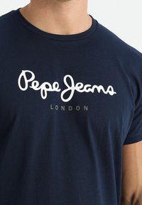 Námořnická modrá tričko nošené osobou, se bílým nápisem "Pepe Jeans" a menším šedým textem "LONDON" pod ním na hrudi.
