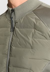 Gevoerde jas in olijfgroen met een ronde kraag, ritssluiting en contrasterende schouderpanelen. Glad oppervlak met gestikte secties.