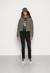 Veste matelassée verte olive cropped avec un col haut, t-shirt noir à motif graphique, jean slim noir et baskets blanches avec des accents noirs.