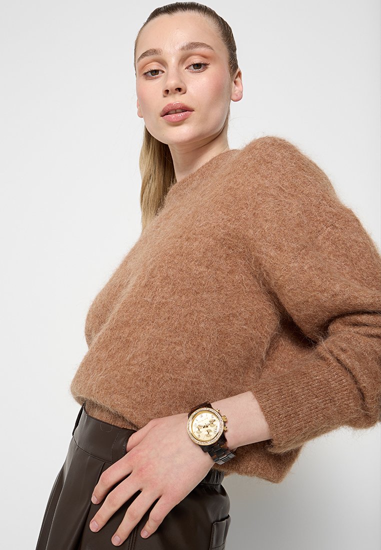 Pull en laine marron et duveteux avec une coupe décontractée, porté sur un pantalon en cuir noir, accompagné d'une montre dorée et marron avec un cadran rond.