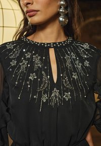 Rochie neagră cu decolteu în formă de cheie, embellishată cu mărgele și paiete argintii în modele de stele și lună, având mâneci transparente.