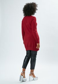 Rote gestrickte Tunika mit langen Ärmeln und geripptem Saum. Kombiniert mit hoch taillierten schwarzen Kunstlederhosen und silbernen Plateau-Absätzen.