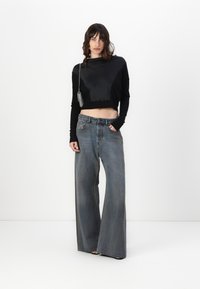 Top corto nero con pannello anteriore in satin, indossato con jeans grigi sbiaditi a vita alta e gamba larga. La modella posa con una mano in tasca.