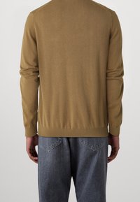 Homme portant un pull à manches longues marron clair et un jean bleu délavé ample, vu de dos sur un fond uni.