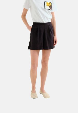 Persona con pantalones cortos plisados negros, camiseta blanca con parche de flor amarilla y zapatos beige sin cordones, de pie con una mano en el bolsillo.