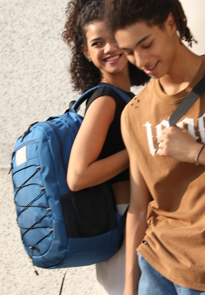 Zaino blu con finitura testurizzata, caratterizzato da un design con corda bungee e una tasca laterale in rete. Due persone vestite in modo casual sullo sfondo.