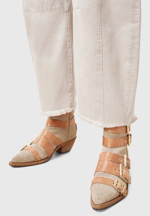 Gros plan sur un pantalon court couleur crème aux ourlets effilochés, porté avec des chaussures pointues en cuir beige et havane, ornées de plusieurs brides à boucles.