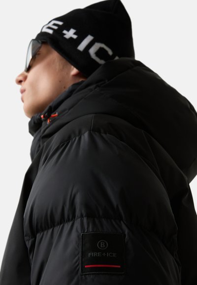 Person in einer schwarzen gepolsterten Jacke mit "Fire+Ice"-Logo auf dem Ärmel, schwarzer Mütze mit weißem Text und großen schwarzen Sonnenbrillen.