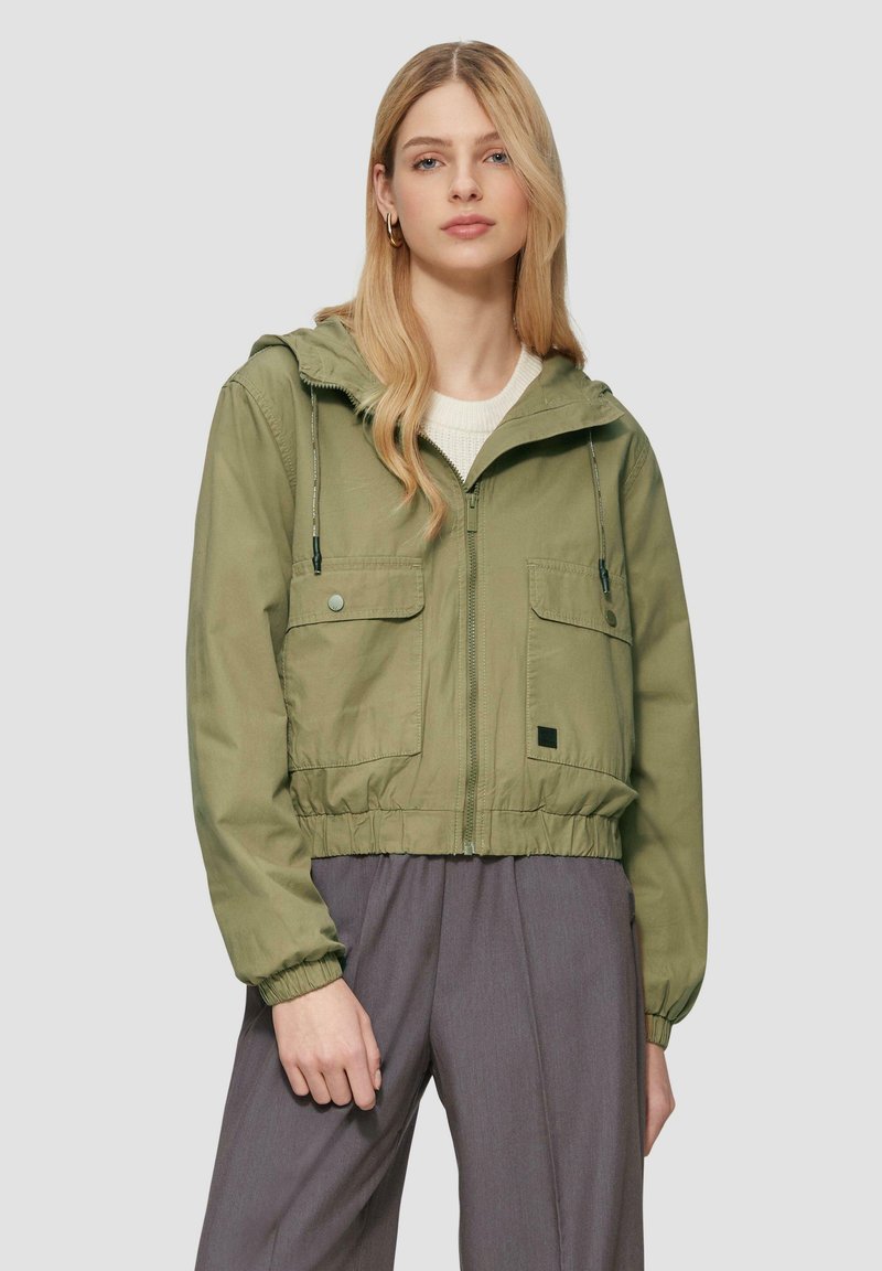 Giacca leggera verde con cappuccio, polsini ed elastico in vita, due tasche frontali e zip frontale. Indossata sopra un top in maglia leggera.
