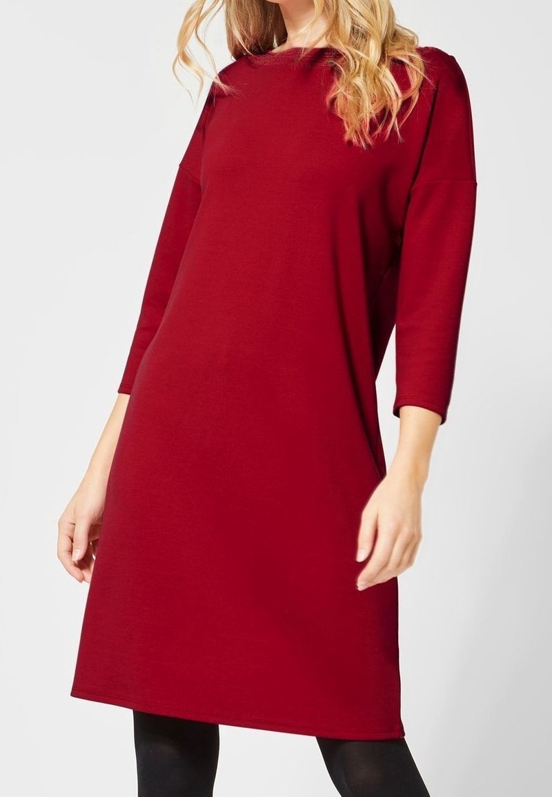 Femme portant une robe rouge mi-longue à manches longues avec un col rond, associée à des leggings noirs sur un fond uni.