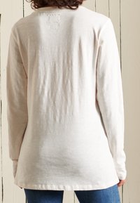 Ljusbeige långärmad skjorta i ett mjukt, texturerat tyg. Har rundad fåll och en liten logotypdetalj vid baksidan av kragen.