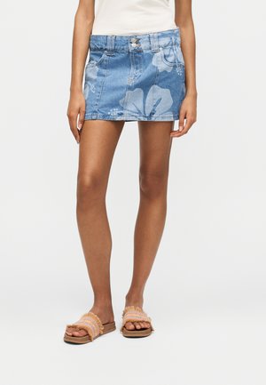 Femme portant une mini-jupe en jean bleue avec des motifs floraux, un haut blanc et des sandales à franges, debout devant un fond uni.