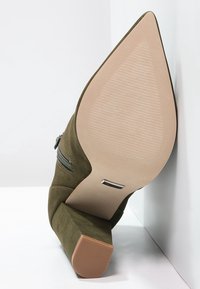 Botín de ante verde con tacón y punta afilada, detalle de cremallera en el lateral y una suela beige lisa con finas líneas de agarre texturizadas.