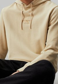 Sudadera con capucha beige hecha de algodón, que cuenta con un bolsillo frontal, cordones y un logo en relieve en el pecho. Textura suave, ajuste relajado.