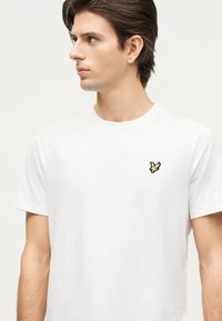 Νέος άνδρας με σκούρα μαλλιά που φοράει απλό λευκό t-shirt με μικρό κίτρινο πουλί στο αριστερό στήθος, κοιτάζει προς τα αριστερά.