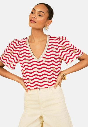 Woman wearing a red and white wavy striped blouse with gold sun patterns, beige pants, sun-shaped earrings, and gold bracelets, hands on hips.