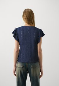 Mørkeblå topp med korte flate ermer, med et strukturert mønster og utklippsdetaljer. Kombinert med falmede denimbukser.