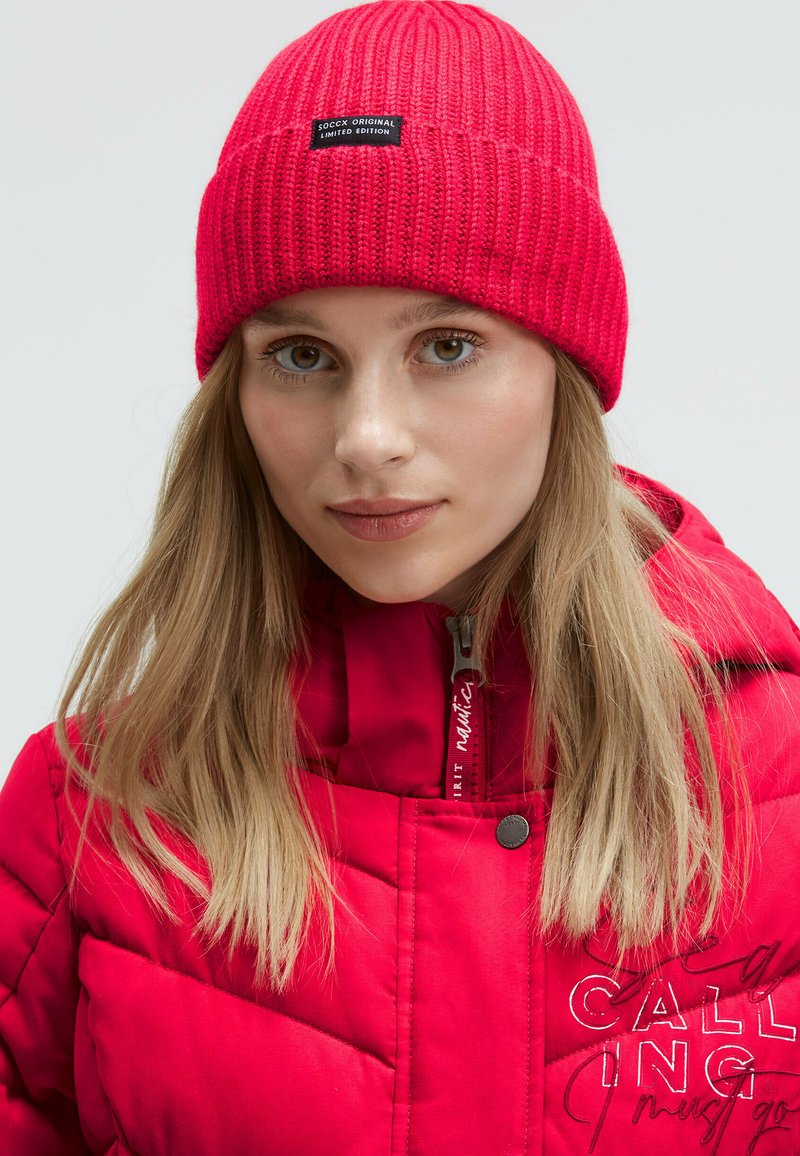 Red knitted beanie with ribbed texture, featuring a label. Red puffy jacket with quilted pattern and embroidered text in contrasting colors.