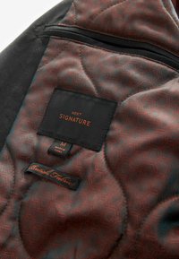 Juodas siuvinėjimo audinys, kuriame yra šešiakampė raštas tamsiai raudona. Apima etiketes: "NEXT," "SIGNATURE," ir "British Fabric."