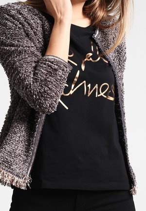 Kobieta ubrana w szarą, teksturowaną kurtkę na zamek, na czarnej koszulce z błyszczącym, złotym napisem w stylu kursywy z przodu.