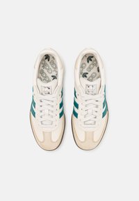 White athletic shoes with teal stripes, suede accents, and a textured sole. Features logo on tongue and patterned insole. Laced up.