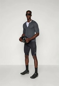 Kortærmet grå cykeltrøje med lynlås, kombineret med sorte shorts. Modellen holder et cykelhjelm og bærer sorte solbriller og cykelsko.