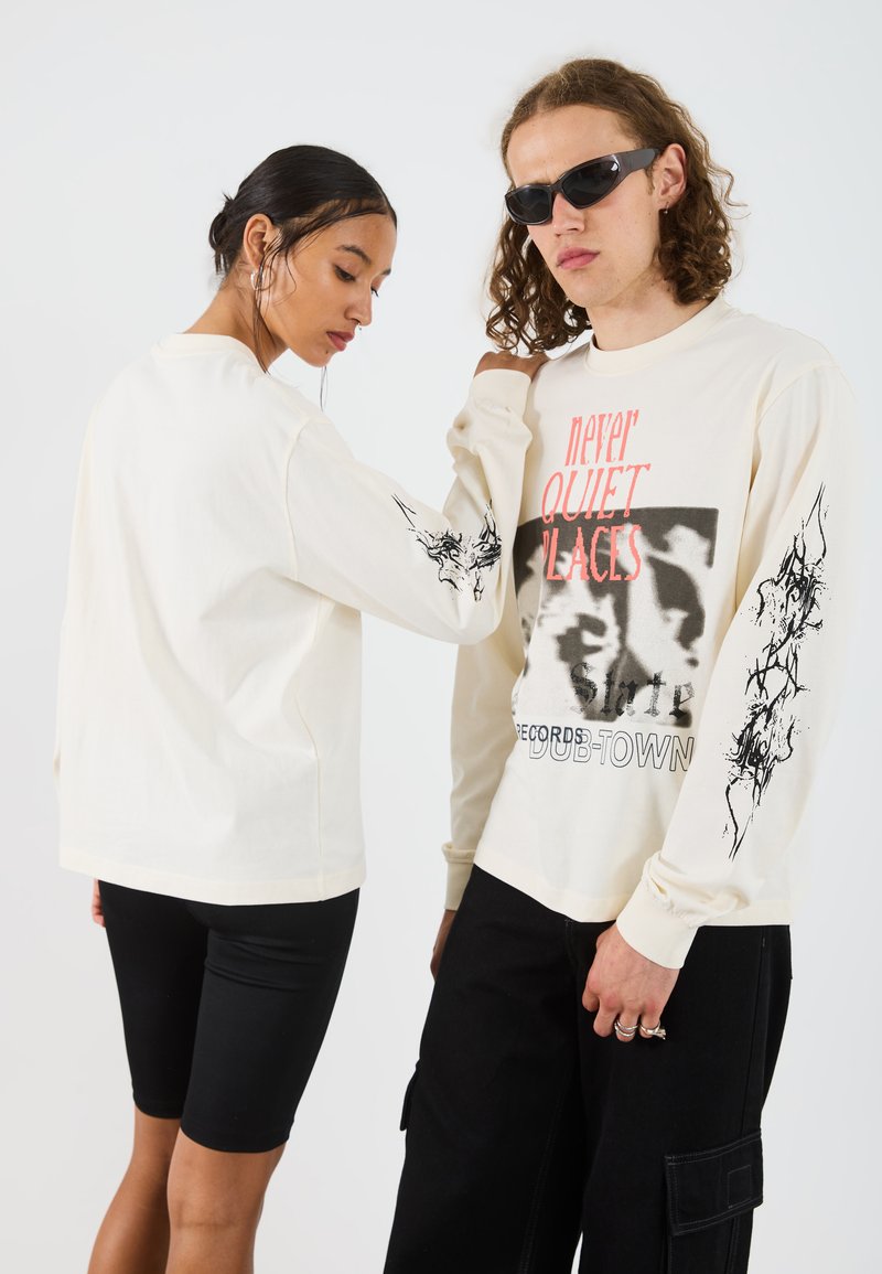 Long cream t-shirt featuring a graphic print and text on the front, with black ink designs on the sleeves. Styled with black leggings.