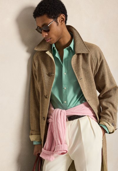 Mand iført brun fiskeben-frakke, grøn skjorte med knapper, hvide bukser, lyserød sweater bundet om taljen og mørke solbriller.