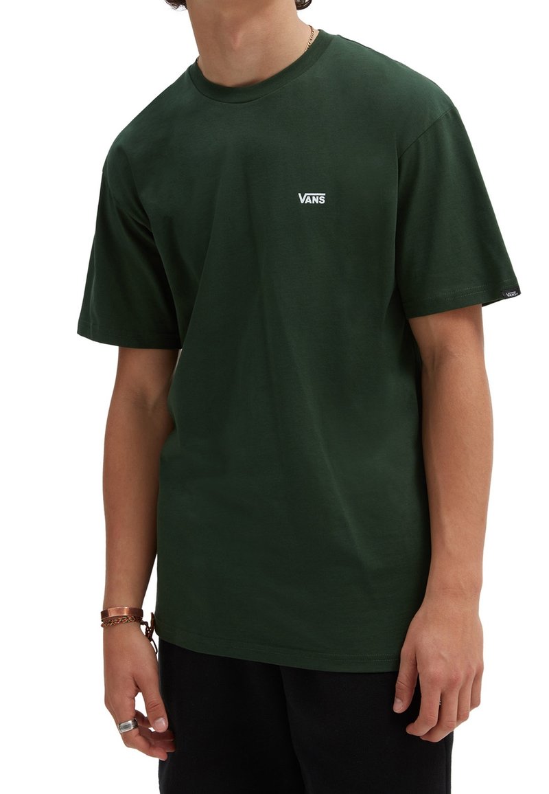 Personne portant un t-shirt Vans vert foncé à manches courtes avec un petit logo blanc sur la poitrine et une étiquette logo assortie sur la manche gauche.