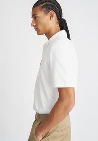 Witte polo met korte mouwen en een kraag, met een klein logo op de borst. Gekleed met beige broeken, weergegeven in zijaanzicht.