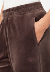 Brūni, mīksti auduma sporta bikses ar elastīgu jostasvīru, sānu kabatām un mazu iedziļinātu logotipu uz kreisā augšstilba.