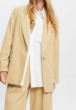 Osoba o blond włosach ubrana w beżową sztruksową marynarkę i pasujące spodnie, pod którymi ma białą koszulę na guziki, na jednolitym tle.