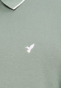 Ανοιχτό πράσινο υφασμάτινο ύφασμα με μικρό λευκό κεντητό πουλί κοντά στο πάνω κέντρο και μέρος διπλωμένου γιακά.