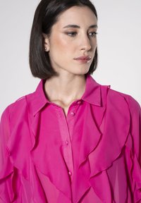 Blusa fucsia con strati arricciati, maniche lunghe e chiusura a bottoni. Realizzata in un tessuto leggero e trasparente con colletto classico.