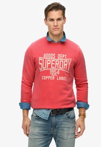 Roter Baumwollpullover mit weißem "SUPERDRY"-Text, Rundhalsausschnitt und langen Ärmeln, über einem hellblauen Denimhemd getragen. Kombiniert mit hellen Denimjeans.