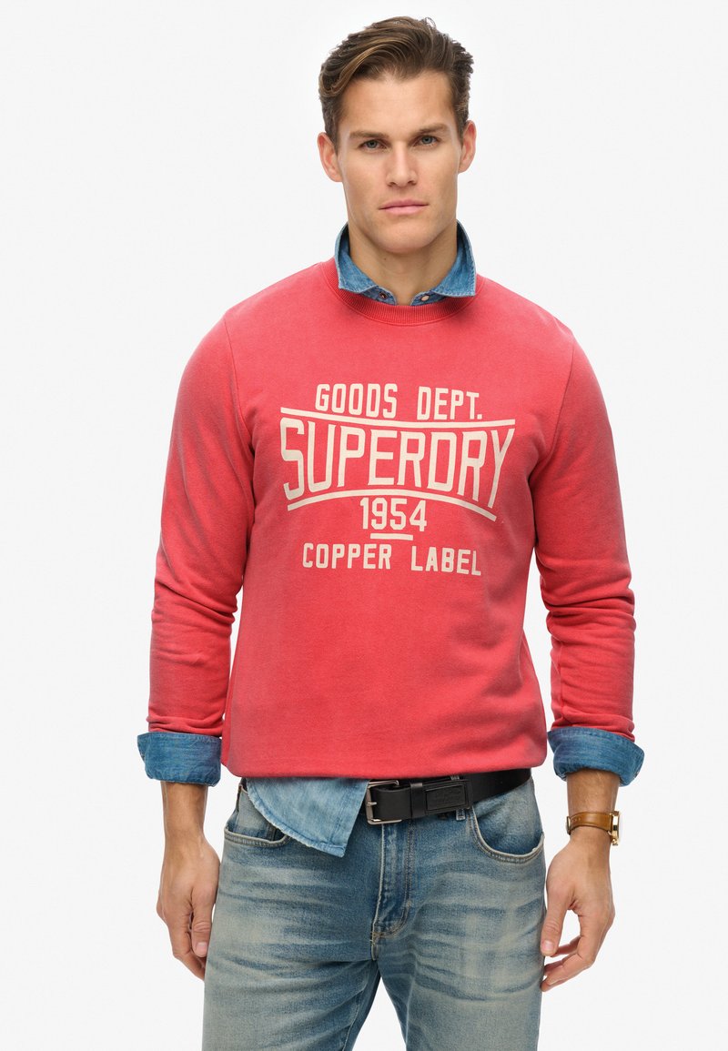 Roter Baumwollpullover mit weißem "SUPERDRY"-Text, Rundhalsausschnitt und langen Ärmeln, über einem hellblauen Denimhemd getragen. Kombiniert mit hellen Denimjeans.