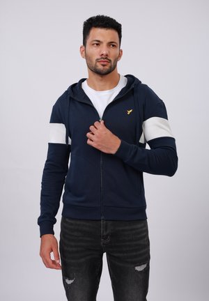 Navy blue zip-up hoodie with white shoulder stripes, kangaroo pocket, and gold logo. Worn with a white t-shirt and distressed black jeans.