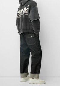 Sudadera negra de gran tamaño con texto blanco y mangas deshilachadas, combinada con pantalones de mezclilla oscuros con puños doblados y bolsillos laterales. Zapatillas blancas.